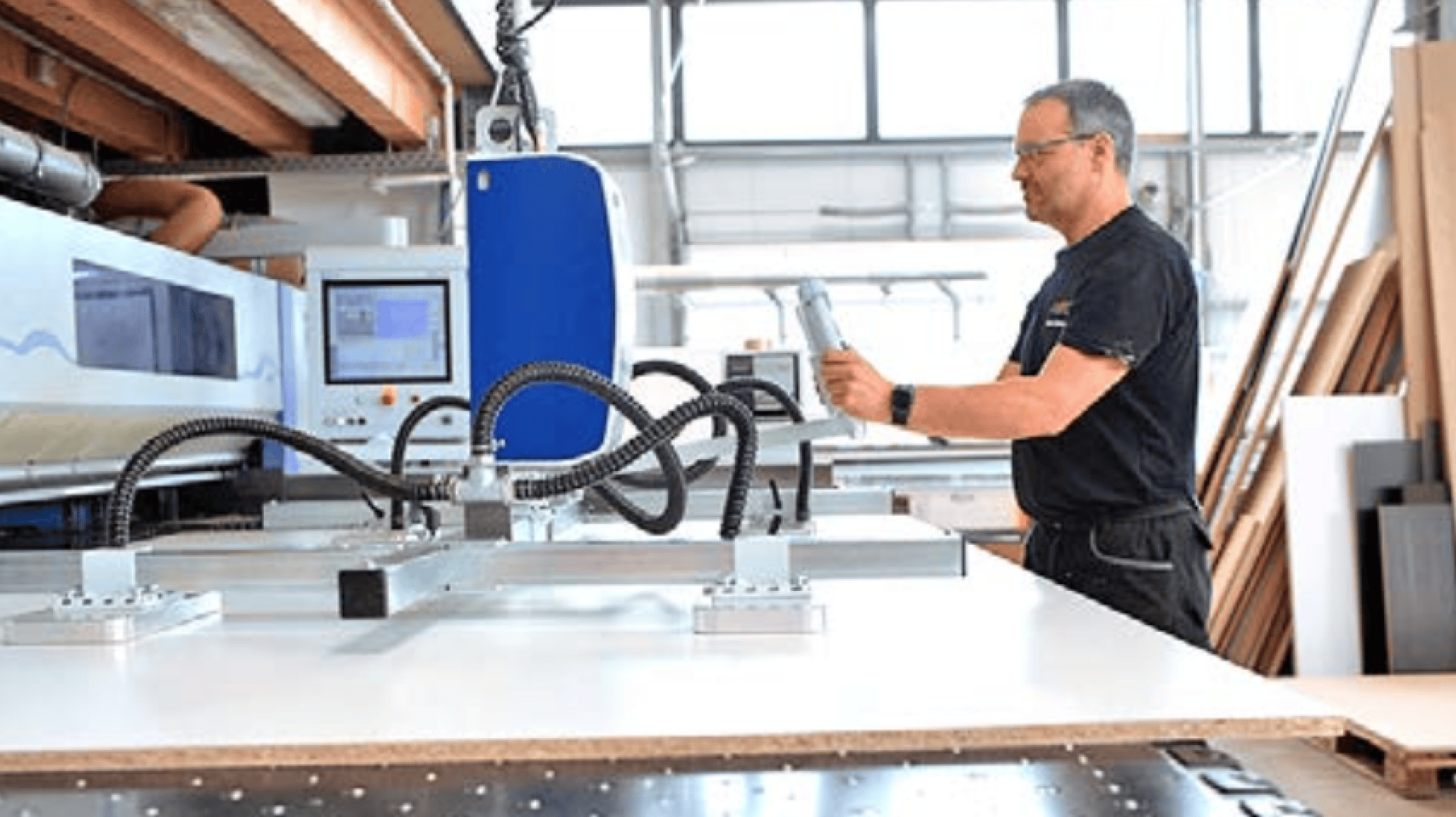 Ein Mann bedient eine große Industriemaschine in einer hellen, modernen Werkstatt und prüft ein Tablet oder ein Steuergerät in der Nähe einer flachen weißen Tafel auf der Maschine.